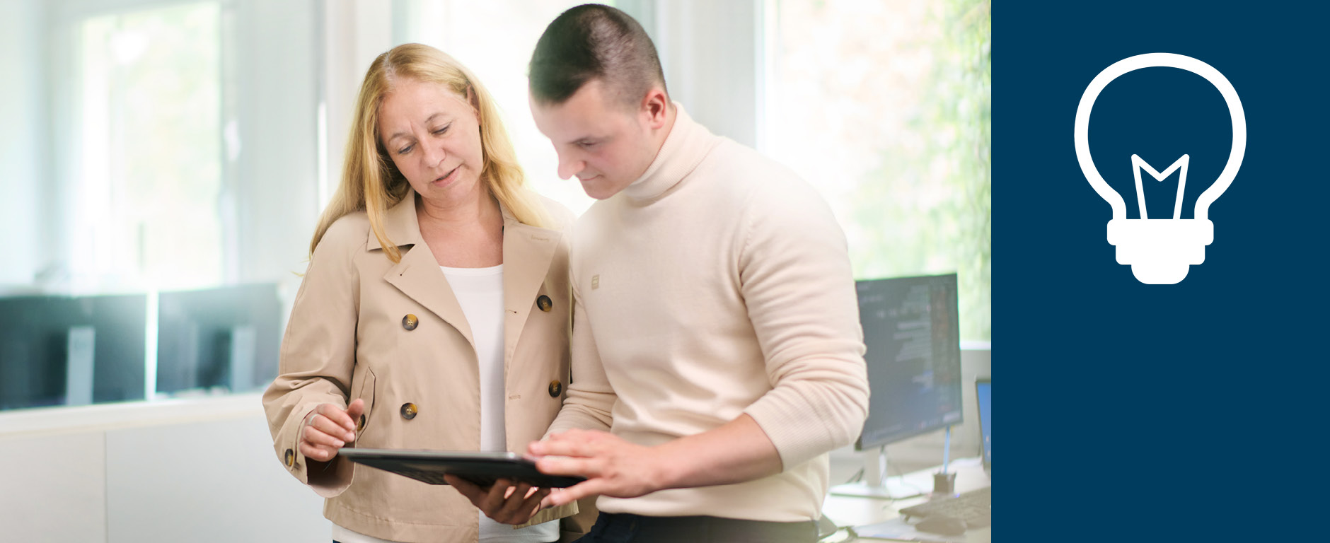 Eine Frau und ein Mann im Büro stehen nebeneinander und schauen gemeinsam auf ein Tablet.